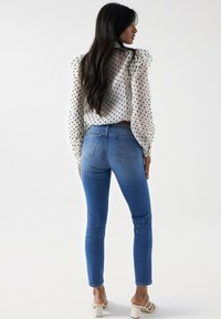 Jean bleu skinny avec une taille mi-haute et des poches arrière, associé à un chemisier blanc à pois noirs et manches bouffantes.
