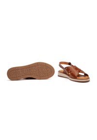 Sandalia de cuero marrón con punta abierta, correas anchas con detalles metálicos y suela de goma beige con un fondo estampado.