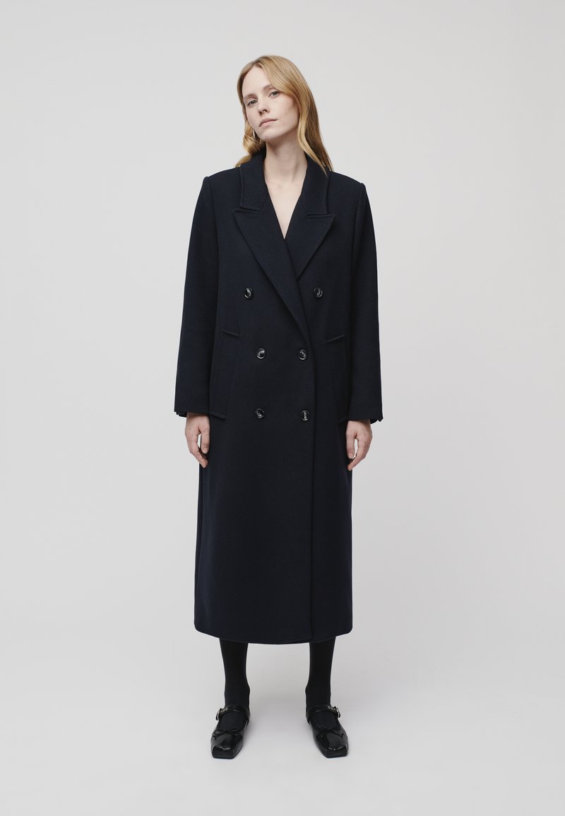 Dark navy double-breasted wool coat with large lapels, front pockets, and black buttons. Mid-calf length, straight silhouette, and long sleeves.