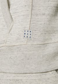 Hoodie en polaire grise avec un tissu texturé, présentant un ourlet et des poignets côtelés. Inclut une étiquette avec "DNM WRKRS" en lettres bleues.