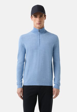 Hellblauer Zip-Up-Pullover aus weichem Strickstoff, mit hohem Kragen und langen Ärmeln, kombiniert mit dunklen Hosen.
