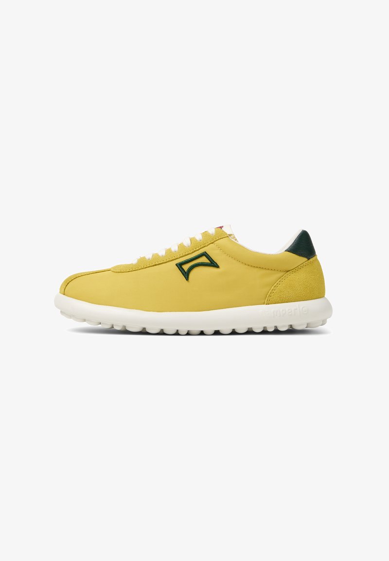 Zapatilla amarilla con parte superior de textil suave, suela de goma blanca y acentos verdes en el talón y el logo. Presenta cordones blancos y una forma redondeada.