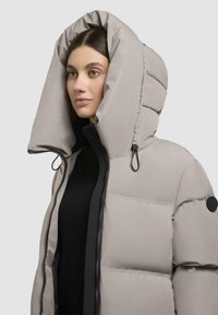 Puffer jacket in light gray with a large, structured hood. Features a black zipper and drawstring accents. Smooth, padded texture.