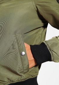 Olive green bomber jacket o gładkiej, błyszczącej powierzchni, z metalowym zatrzaskiem na kieszeni i czarnymi ściągaczami.