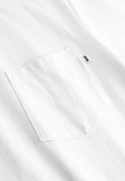 Hvitt stoff med en sydd firkantet lomme som har en liten hvit tagg med svart tekst på øvre høyre kant av lommen.