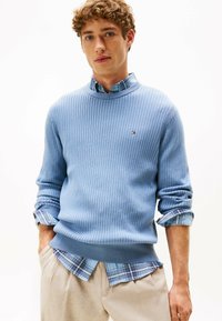 Młody mężczyzna w jasnoniebieskim swetrze prążkowanym, noszący niebieską koszulę w kratę z podwiniętymi rękawami i beżowe spodnie, stoi z rękami w kieszeniach.