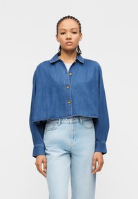 Νεαρό άτομο με πλεξούδες, φοράει κοντή μπλε τζιν ζακέτα με κουμπιά και φαρδιά τζιν παντελόνια με υψηλή μέση, σε λευκό φόντο.