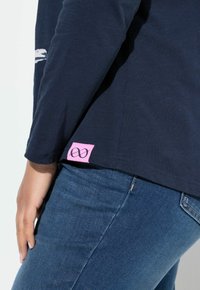 Marineblaues Langarmshirt aus weichem Stoff mit einem rosa Etikett am Saum, das ein Unendlichkeitssymbol zeigt. Kombiniert mit dunkelblauen Jeans.