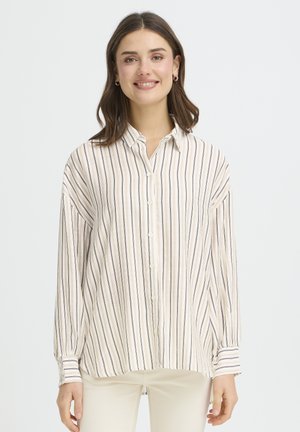 Femme aux cheveux bruns mi-longs portant des boucles d'oreilles créoles dorées et une chemise crème à boutons avec des rayures verticales noires et beige.