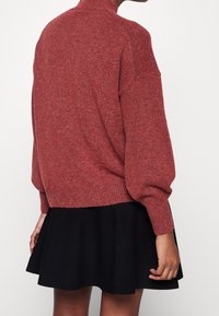 Persona vestita con un ampio maglione rosso a maglia e una gonna nera a campana, vista di spalle su uno sfondo chiaro e uniforme.