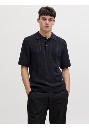 Jeune homme aux cheveux courts et foncés, portant un polo en maille noire à manches courtes et un pantalon noir, debout les mains dans les poches.