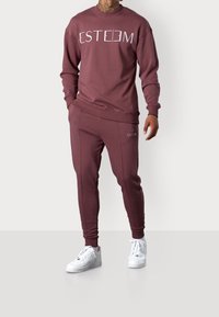 Felpa color marrone e pantaloni sportivi con testo bianco "ESTEEM". Tessuto liscio con polsini e fascia in vita a costine. Abbinato a sneakers bianche.