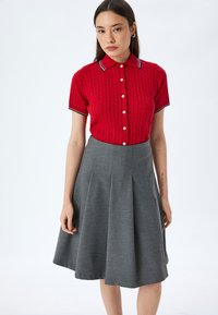 Polo rosso a maglia con maniche corte e bottoni bianchi, abbinato a una gonna grigia svasata con pieghe. Colletto bianco con dettagli blu navy.