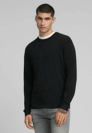 Pull noir à manches longues, avec poignets et ourlet côtelés, col rond orné d'un accent blanc, confectionné en tissu doux, avec un petit logo sur la poitrine.