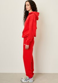 Sarkans vilnas kapucis džemperis ar saskaņotām treniņbiksēm, uz kurām ir neliels balts logo, valkāts ar pelēkiem un ziliem sporta apaviem.