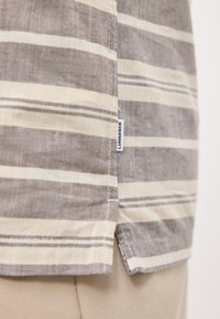 Μπεζ και γκρι ριγέ λινό πουκάμισο με μικρή ετικέτα μάρκας "Lindbergh" φορεμένο πάνω από ανοιχτό μπεζ παντελόνι.