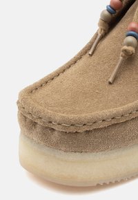 Mocassim de camurça castanho-claro com bico arredondado, sola de borracha bege e atacadores decorativos em pérolas com detalhes em castanho, azul e rosa.