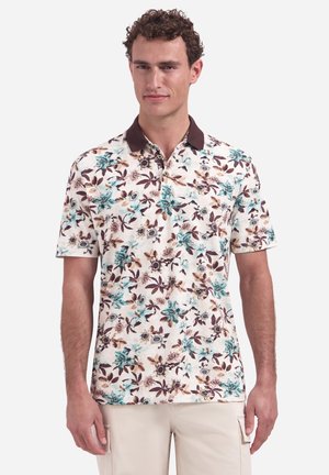 Mann trägt ein kurzärmeliges, florales Polohemd mit braunem Kragen und hellbeige Cargohosen, steht vor einem einfarbigen Hintergrund.