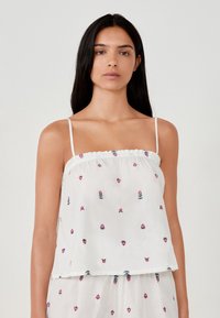 Kobieta z długimi ciemnymi włosami, ubrana w białą, bezrękawnikową bluzkę i pasujące do niej spodenki z małym różowym i zielonym wzorem kwiatowym.