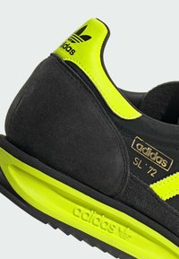 Černé semišové tenisky se žlutými akcenty, s vysokou podrážkou a zlatým brandingem. Design zahrnuje retro pruhy a polstrovaný límec.