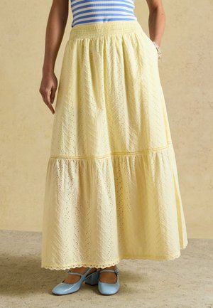Femme portant une jupe midi jaune ajourée, un haut rayé bleu et blanc, et des chaussures Mary Jane bleu clair, debout sur un sol beige.
