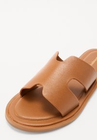 Braune Sandale mit einem breiten Riemen und Ausschnittdetails, aus glattem synthetischem Material gefertigt. Flacher Boden mit abgerundeten Kanten.