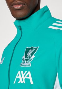 Teal lynlåsjakke med hvide striber, Liverpool Fodboldklubs logo og AXA-branding, med en glat, let tekstur.
