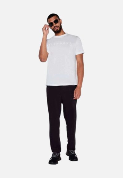 Camiseta blanca de manga corta con un logo sutil, combinada con pantalones negros y zapatillas negras. La persona lleva gafas de sol oscuras.