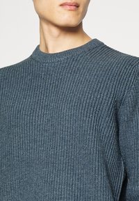 Homme portant un pull en maille côtelée bleu, coupé sous le nez, sur un fond blanc.