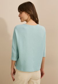 Ljusblå, texturerad tröja med tre fjärdedelsärmar, rund halsringning och en något lös passform. Baksidan har en ren sömdetalj.