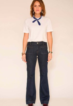 Jean évasé en denim foncé avec poches avant et arrière, associé à un t-shirt blanc et un foulard bleu à motifs autour du cou. Texture lisse et coupe classique.