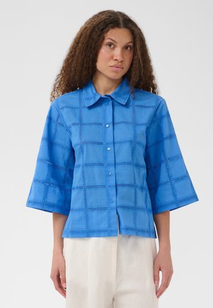 Femme aux cheveux bouclés portant un chemisier bleu vif à manches courtes boutonné avec un motif ajouré à carreaux et un pantalon blanc.