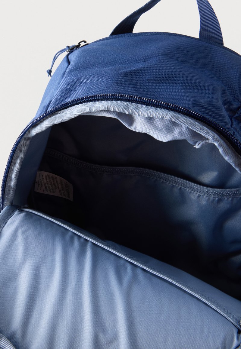 Blauer Rucksack mit offenem oberen Reißverschluss, der das leere Hauptfach und die Innentasche mit Stoffetikett im Inneren zeigt.
