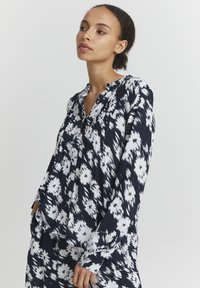 Chemise à manches longues bleu marine et blanche à motifs, avec col mandarin, coupe décontractée et des détails de boutons, présentant des motifs floraux et des plis.