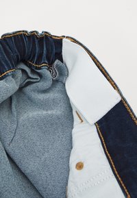 Modra denimska pasnica z elastiko, bela notranja podloga z bež šivi in dvema rjavima gumboma.