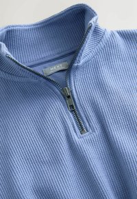 Pullover azzurro chiaro testurizzato con colletto alto e chiusura a zip in argento. Presenta un sottile motivo a coste e un'etichetta con marchio all'interno.