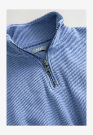 Jasnoniebieski, teksturowany pulower z wysokim kołnierzem i srebrnym zamkiem błyskawicznym. Posiada subtelny, prążkowany wzór oraz metkę z logo wewnątrz.