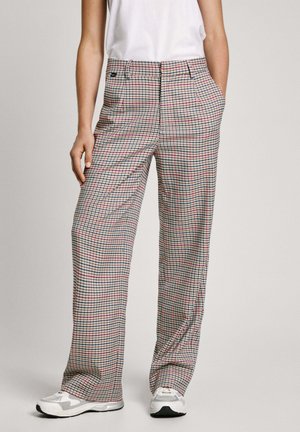 Femme portant un pantalon taille haute à jambes larges à carreaux multicolores, des baskets blanches et un débardeur blanc, debout les mains dans les poches.