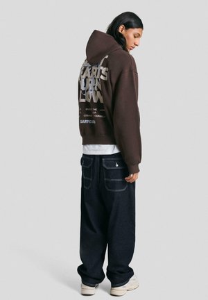 Rjava kapuca s kapuco in grafičnim besedilom na hrbtni strani, v kombinaciji s temno jeans baggy hlačami in belimi supergami. Gladka tekstura in sproščen kroj.