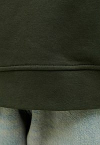 Sweter w oliwkowym kolorze z wykończeniem w teksturowany, prążkowany sposób, w zestawie z jasnoniebieskimi dżinsami w lekko wyśmierzwionym wzorze.