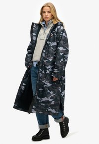 Kurtka przeciwdeszczowa w kamuflażu w ciemnoszarym i niebieskim odcieniu, o długim kroju i dużych kieszeniach z przodu. Noszona na szarym hoodie i dżinsach.
