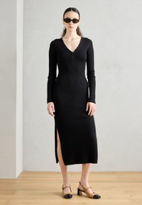 Long black ribbed dress with a V-neck and button detail, featuring long sleeves and a high side slit. Paired with brown and black block-heeled shoes.