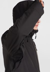 Chaqueta negra resistente a la intemperie con capucha, cierre frontal con cremallera, paneles laterales de malla y cuerdas ajustables. Textura suave y diseño estructurado.