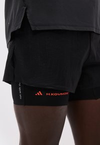 Gros plan sur un short de course noir à couches superposées avec tissu perforé et les logos rouges Adidas et H.Koumori sur une peau foncée.