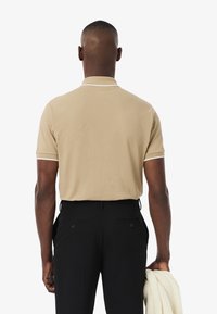 Beige polo skjorta med vit piping på krage och ärmar, tillsammans med svarta byxor. Slät textur, korta ärmar och minimal design.