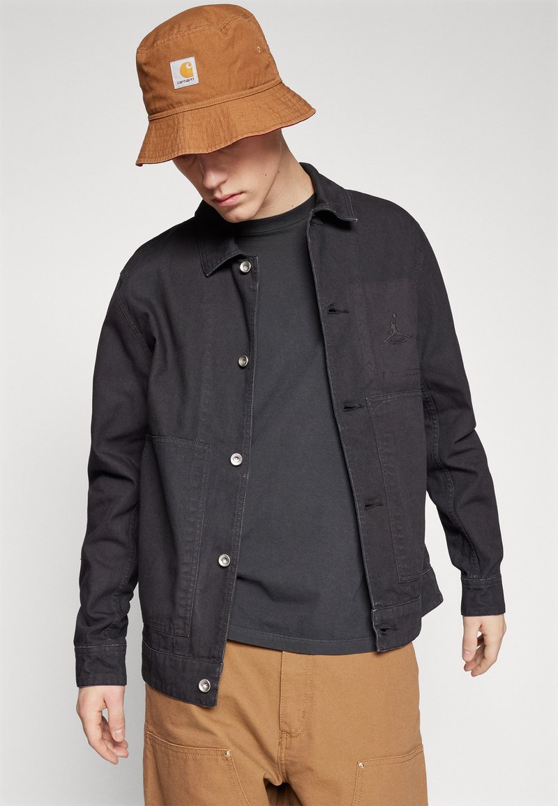 Młody mężczyzna w brązowej czapce typu bucket od Carhartt, czarnej zapinanej kurtce, czarnej koszuli i brązowych spodniach, patrzący w dół na jednolitym tle.