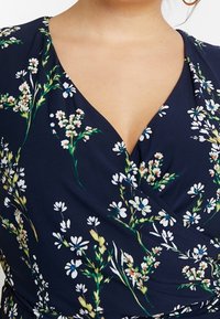 Marinblå omlottskärning klänning med blommönster som har vita prästkragar och gröna stjälkar. Slät tyg med djup V-ringning och veckad midjedetalj.