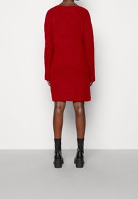 Robe tricotée rouge à manches longues, coupe fluide, tissu texturé, tombant au-dessus des genoux. Associée à des bottines chunky noires à la cheville.