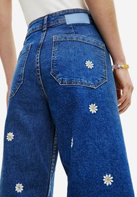 Denim wide-leg pants with a deep blue color, featuring floral embroidery accents. Distinctive back pockets and a branded label at the waist.