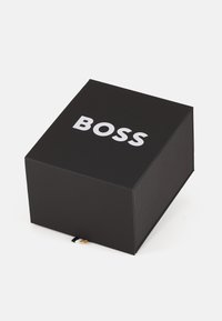 Boîte rectangulaire noire avec "BOSS" en lettres blanches en gras à l'avant. Texture lisse et mate, avec un petit détail d'étiquette noir et blanc.